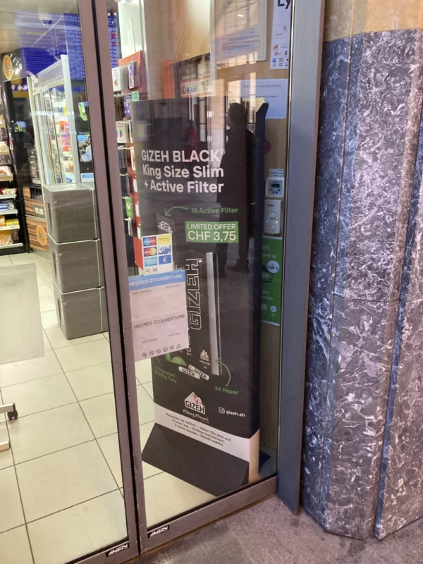 Publicité sur un grand panneau pour du papier à tabac à l'entrée du kiosque de la gare de Lausanne.